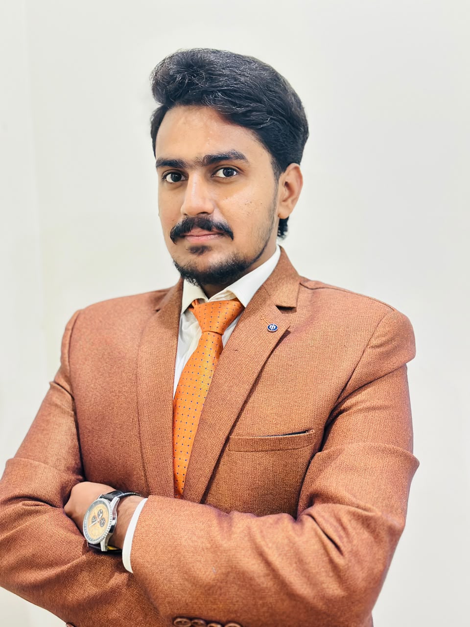 Professional headshot of M. Abdullah Shah, wearing a rust-colored blazer with an orange tie, arms crossed, against a plain light background.